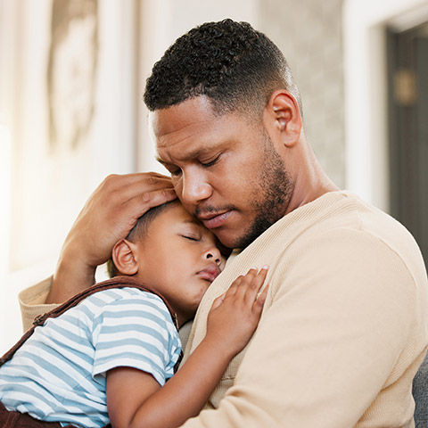 Worried dad hugging his young boy resting on a living room family couch.