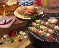 A hamburger with many topping options on a picnic table behind a barbeque grilling shish kabobs