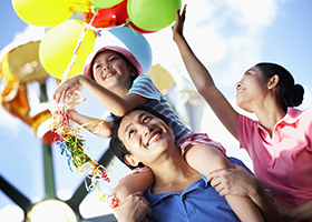 A child sitting on her father's shoulders holding balloons with her mom next to them.